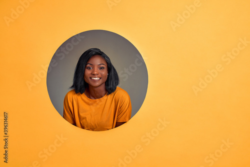 Portrait of cheerful glad  woman with white toothy smile in orange t-shirt looking at camera. Woman in a round hole in the orange wall. Copy space. © HBS