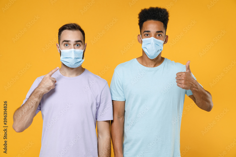 Perplexed young friends european african american men 20s in t-shirts pointing on sterile face mask to safe from coronavirus covid-19 showing thumb up isolated on yellow background studio portrait. Perplexed young friends european african american men 20s in t-shirts pointing on sterile face mask to safe from coronavirus covid-19 showing thumb up isolated on yellow background studio portrait.