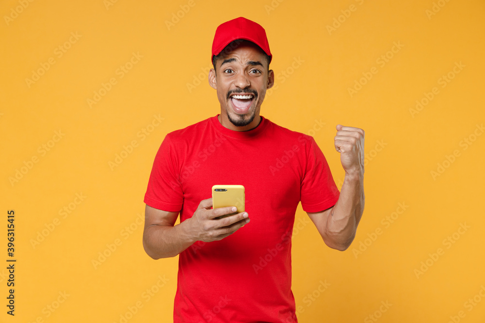 Smiling delivery employee african man 20s in red cap blank print t-shirt uniform workwear work courier dealer service concept hold in hand using mobile cell phone isolated on yellow background studio. Smiling delivery employee african man 20s in red cap blank print t-shirt uniform workwear work courier dealer service concept hold in hand using mobile cell phone isolated on yellow background studio.
