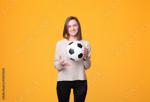 Fototapeta Naklejka Na Ścianę i Meble -  Playful girl smiling at the camera is holding a soccer of football ball over yellow background in a studio.