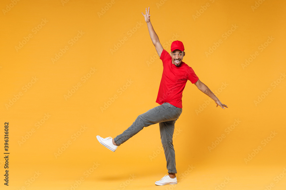 Full length delivery employee african man 20s in red cap blank print t-shirt uniform workwear work courier dealer service during quarantine covid-19 virus concept isolated on yellow background studio.