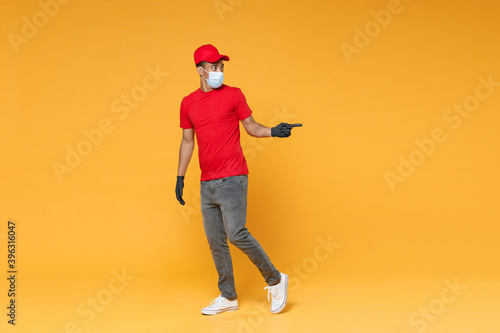 Full length delivery employee african man in red cap t-shirt face mask gloves uniform work courier dealer service on quarantine coronavirus covid-19 virus concept isolated on yellow background studio. © ViDi Studio