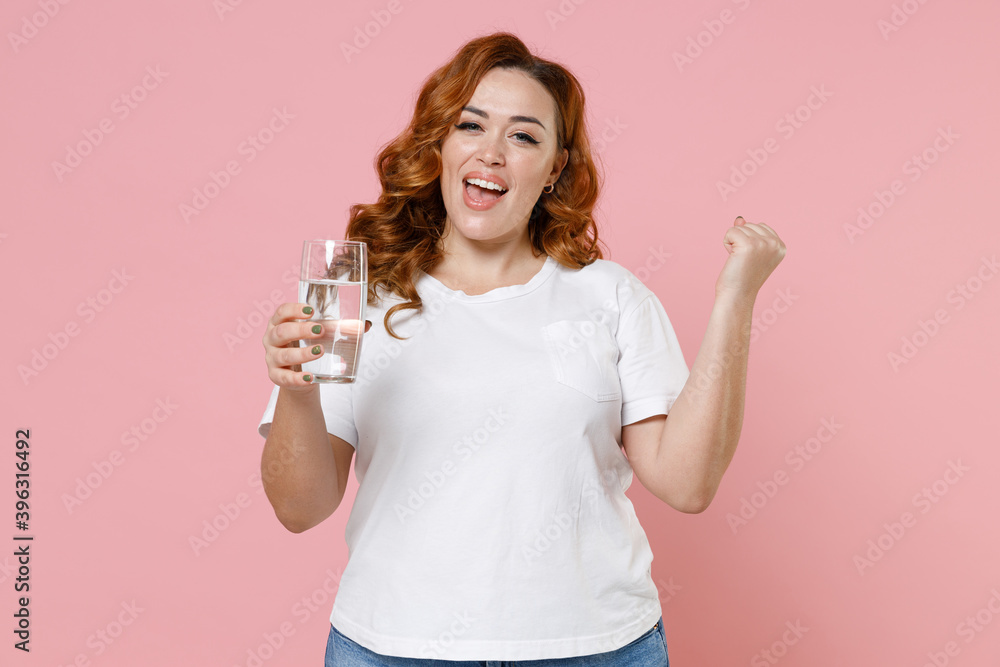 Happy joyful young redhead plus size body positive woman 20s in white casual t-shirt hold glass of water doing winner gesture looking camera isolated on pastel pink color background studio portrait. Happy joyful young redhead plus size body positive woman 20s in white casual t-shirt hold glass of water doing winner gesture looking camera isolated on pastel pink color background studio portrait.
