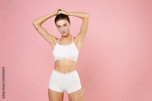 Attractive beautiful fitness young brunette woman 20s in white underwear with perfect body standing posing put hands on head making ponytail isolated on pastel pink colour background, studio portrait.