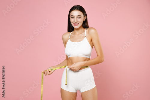 Smiling cheerful thin sports young brunette woman 20s in white underwear posing measuring waist with measure tape looking camera isolated on pastel pink colour wall background, studio portrait. © ViDi Studio