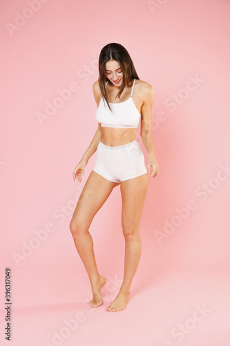 Fototapeta Naklejka Na Ścianę i Meble -  Full length of charming cheerful smiling young brunette woman in white underwear showing fit thin body posing standing looking down isolated on pastel pink colour background, studio portrait.