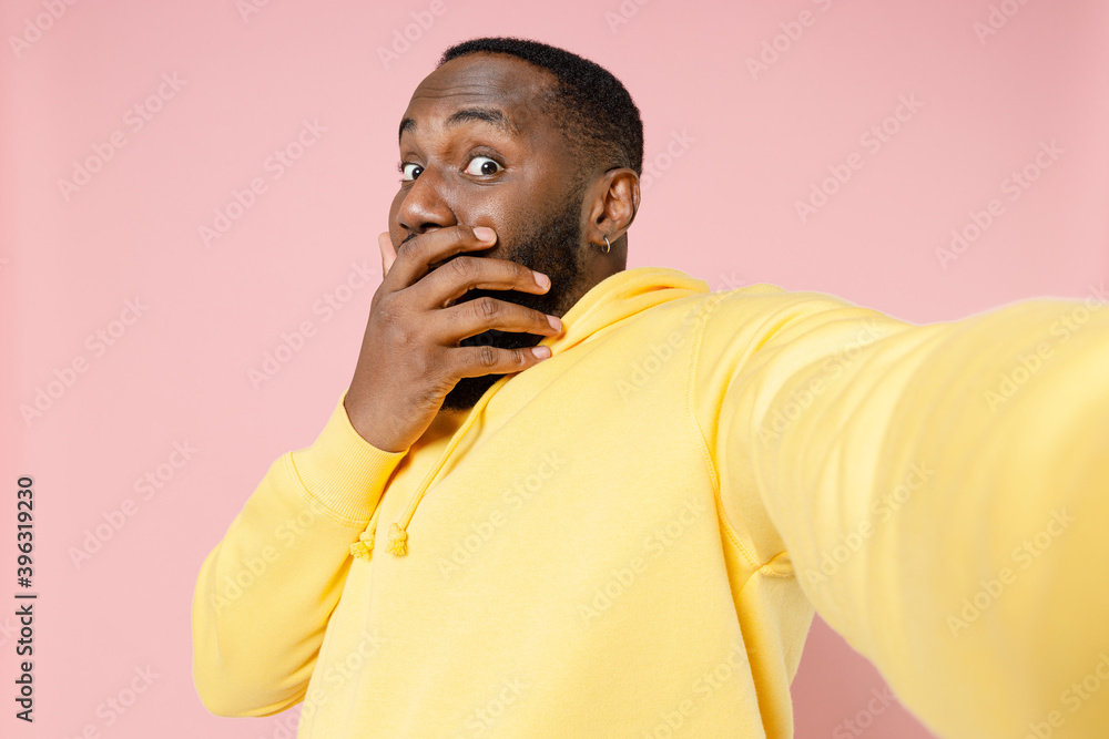 Close up of shocked young african american man in casual basic yellow streetwear hoodie doing selfie shot on mobile phone covering mouth with hand isolated on pastel pink background studio portrait. Close up of shocked young african american man in casual basic yellow streetwear hoodie doing selfie shot on mobile phone covering mouth with hand isolated on pastel pink background studio portrait.