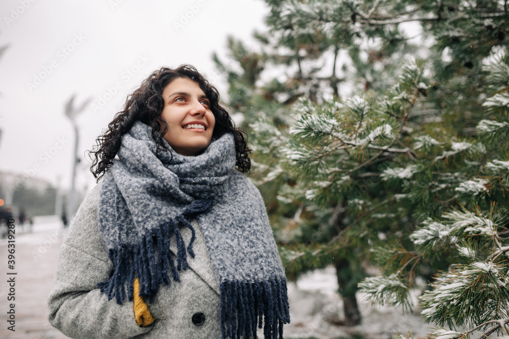 Smiling young woman stands in a frosty snowy winter park. Female wearing a grey coat and blue scarf having fun among fir trees covered with snow. Winter holidays, Christmas and festive mood concept. Smiling young woman stands in a frosty snowy winter park. Female wearing a grey coat and blue scarf having fun among fir trees covered with snow. Winter holidays, Christmas and festive mood concept.