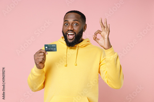 Excited young african american man 20s in casual yellow streetwear hoodie standing hold credit bank card showing OK gesture looking camera isolated on pastel pink color background studio portrait.