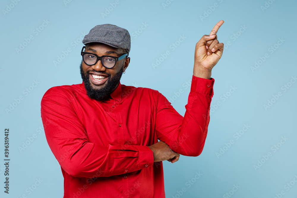 Fototapeta premium Smiling cheerful young bearded african american man 20s wearing casual red shirt eyeglasses cap pointing index finger up on mock up copy space isolated on pastel blue color background studio portrait.