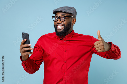 Smiling young bearded african american man 20s in casual red shirt eyeglasses cap showing thumb up using mobile cell phone typing sms message isolated on pastel blue color background studio portrait.