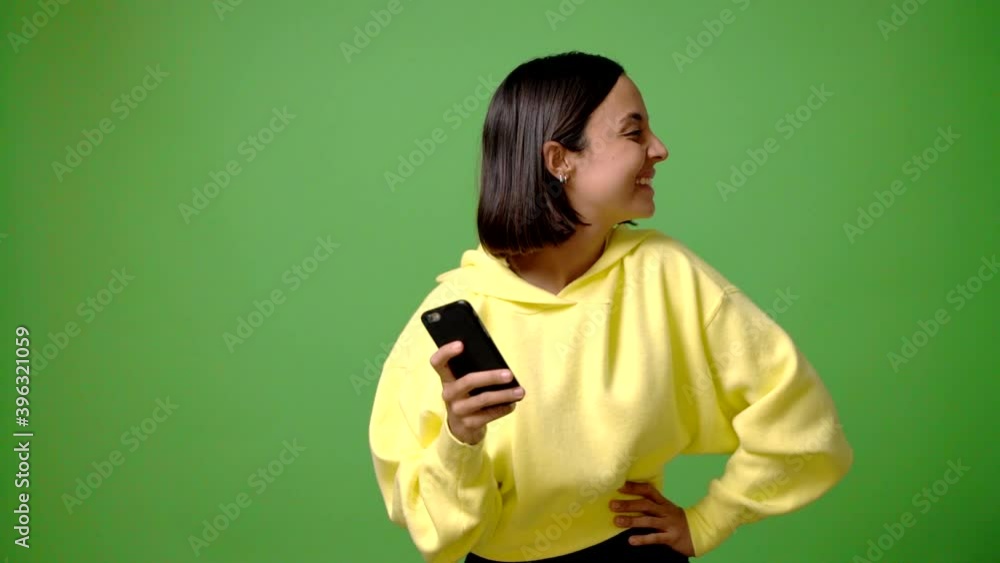 Young woman using mobile phone on green screen chroma key background posing with arms at hip and laughing Young woman using mobile phone on green screen chroma key background posing with arms at hip and laughing