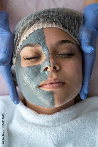 hands of beautician applying beauty mask on young woman face at wellness beauty spa.