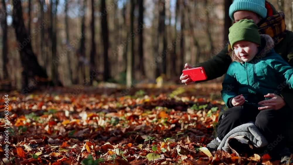 Young stylish mother with little happy laughing boy using smartphone resting in autumn forest enjoying nature beauty on sunny day. Blogging motherhood. Video call. Kids selfie. True feelings. Young stylish mother with little happy laughing boy using smartphone resting in autumn forest enjoying nature beauty on sunny day. Blogging motherhood. Video call. Kids selfie. True feelings.