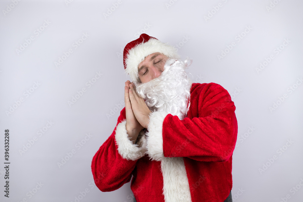 Man dressed as Santa Claus standing over isolated white background sleeping tired dreaming and posing with hands together while smiling with closed eyes. Man dressed as Santa Claus standing over isolated white background sleeping tired dreaming and posing with hands together while smiling with closed eyes.