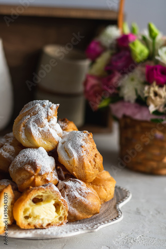 Homemade profiteroles with cream in a gray plate on a white table. Basket with fresh flowers