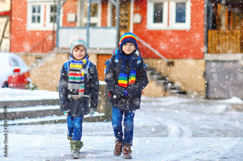 Two little kids boys of elementary class walking to school during snowfall. Happy children having fun and playing with first snow. Siblings ans friends with backpack in colorful winter clothes.