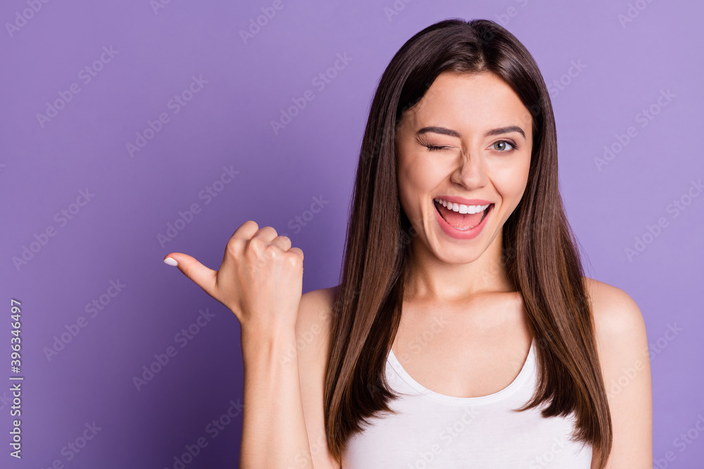 Photo portrait of winking girl pointing thumb at blank space isolated on vivid purple colored background Photo portrait of winking girl pointing thumb at blank space isolated on vivid purple colored background