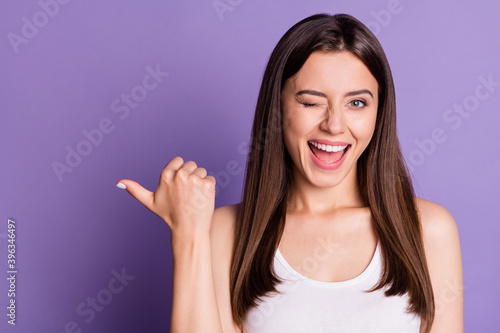 Photo portrait of winking girl pointing thumb at blank space isolated on vivid purple colored background