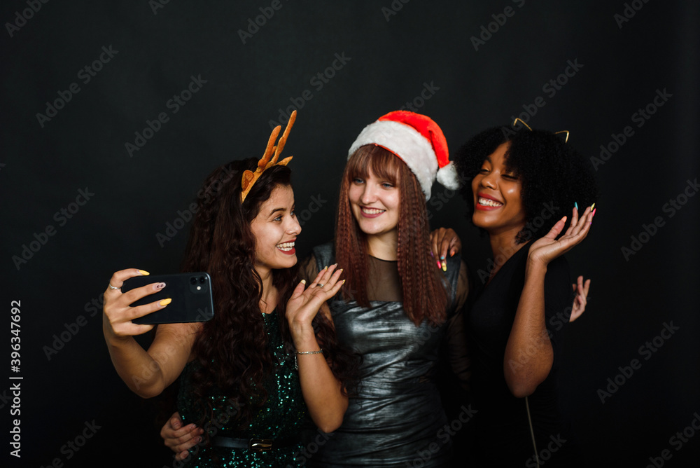 A group of female friends of different nationalities take a Christmas selfie in the studio. Young women in santa hats and deer antlers are having fun together A group of female friends of different nationalities take a Christmas selfie in the studio. Young women in santa hats and deer antlers are having fun together