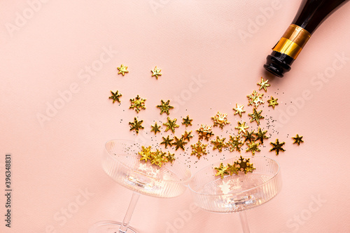 Champagne bottle with golden glittering splashes and champagne glasses on pink background. Flat lay, top view, trendy christmas concept.