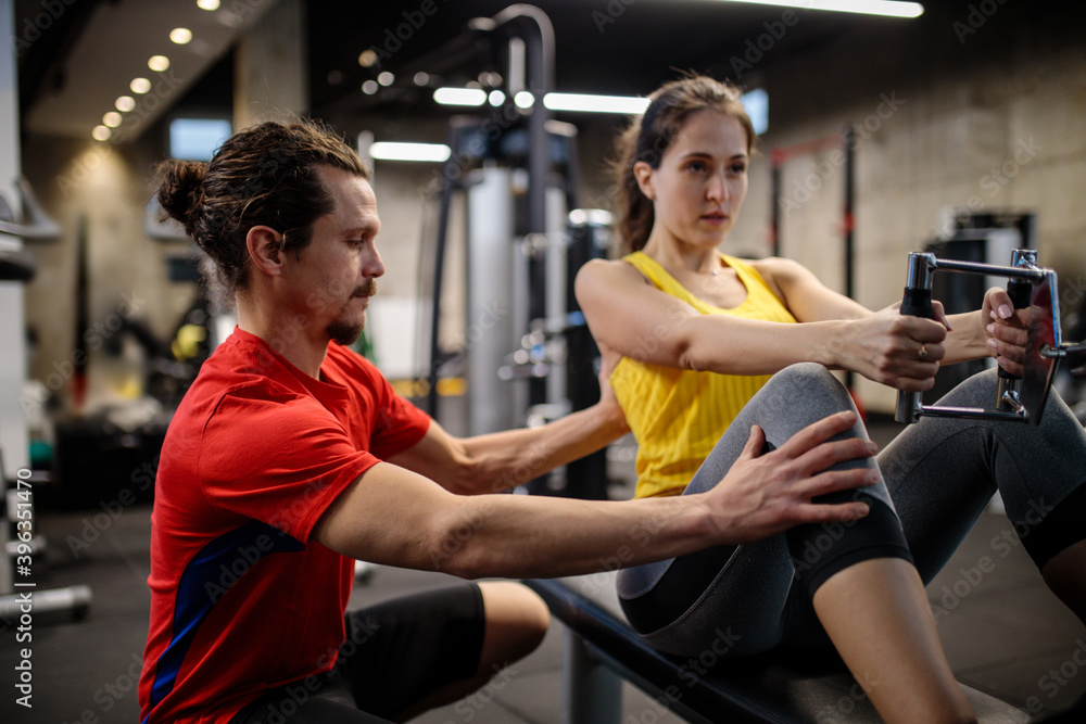 Young Hispanic personal trainer working with young woman who is exercising on a rowing machine in modern indoor gym. Healthy and active sports lifestyle