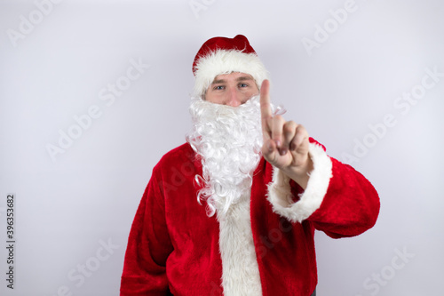 Man dressed as Santa Claus standing over isolated white background showing and pointing up with fingers number one while is serious