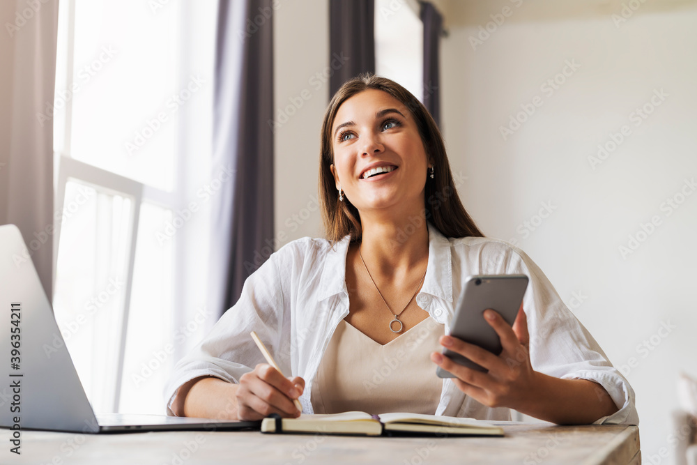 Obraz premium Beautiful brunette girl rejoices uses gadgets. Online training, remote work from home. Business woman sits at table