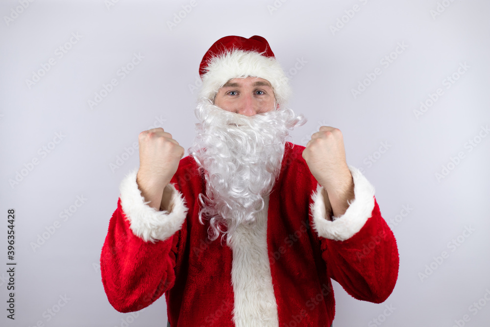 Man dressed as Santa Claus standing over isolated white background very happy and excited making winner gesture with raised arms, smiling and screaming for success. Man dressed as Santa Claus standing over isolated white background very happy and excited making winner gesture with raised arms, smiling and screaming for success.