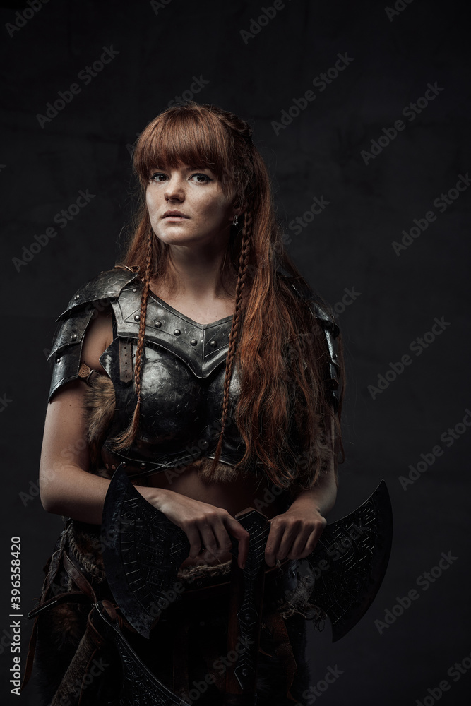 Naklejka premium Beautiful but dangerous warlike woman viking with long brown hairs in dark armour holding huge axe in dark background.