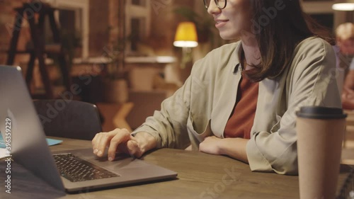 Close up shot of young brunette businesswoman in casualwear sitting at office desk with to go coffee on it, smiling and using touchpad on laptop while browsing the Internet in the evening