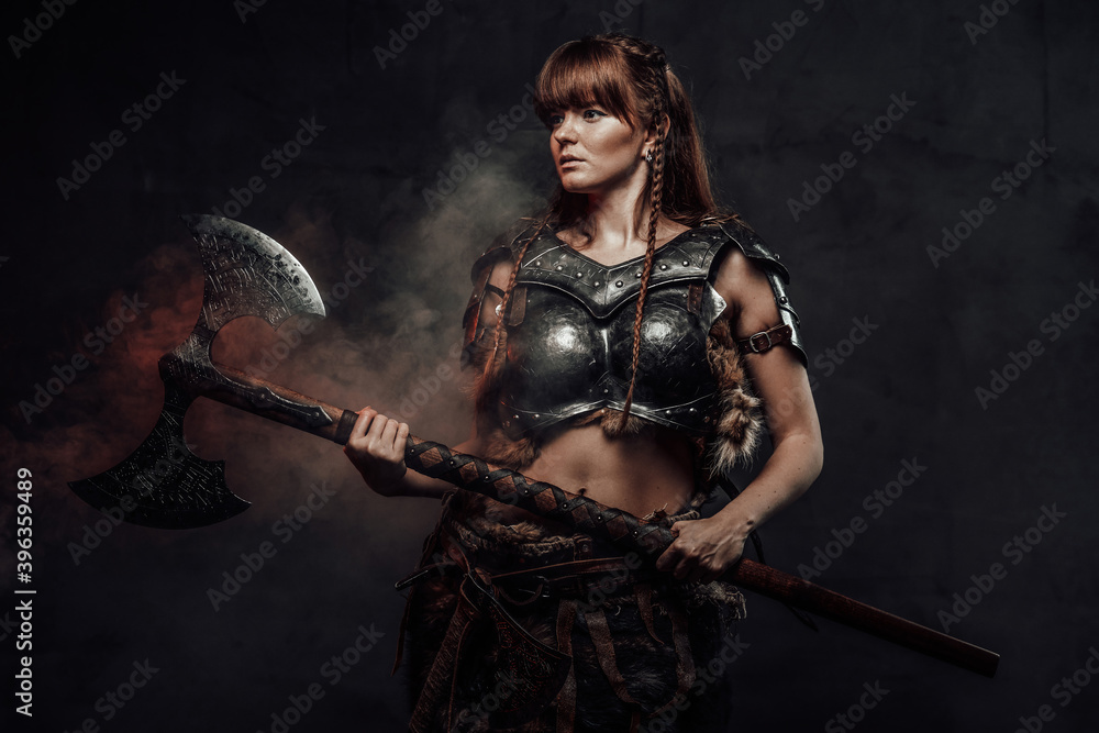 Fototapeta premium Atmospheric portrait of scandinavian amazon with brown hairs dressed in light armour holding two handed axe in dark background.