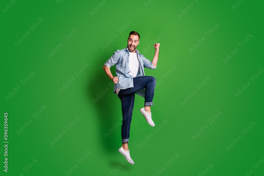 Full size profile photo of impressed funny guy jump wear blue shirt jeans footwear isolated on green color background Full size profile photo of impressed funny guy jump wear blue shirt jeans footwear isolated on green color background