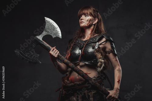 Wielding two handed axe grimy nordic amazon with brown hairs in dark armour posing in dark background.