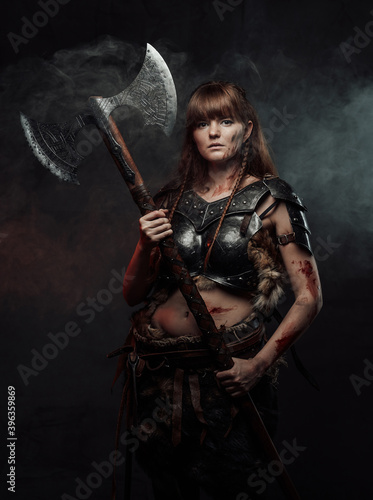Armed with two handed axe nordic grimy amazon in dark armour with brown hairs poses in dark background with smoke. © Fxquadro