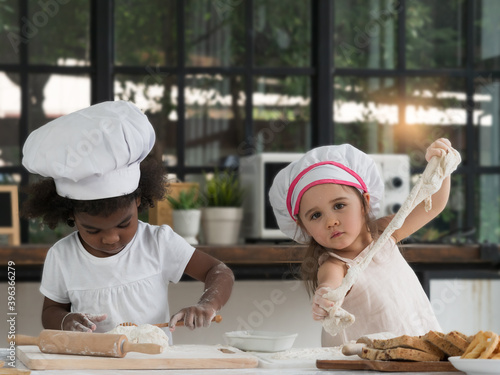 Two cute little diversiy girls wearing chef hat enjoying and helping each other baking and knead the dough. Diverse and Education concept. photo