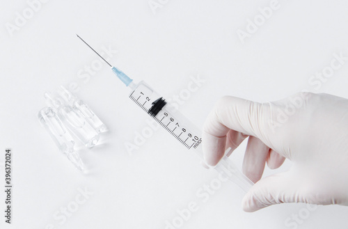 A hand in a white medical glove holds a syringe. In the background, medicine in vials.
