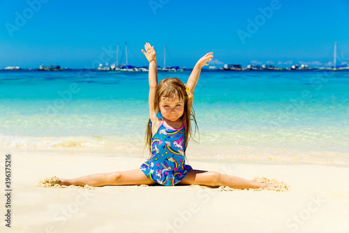 Smiling little girl doing gymnastics splits on a sandy tropical beach. The concept of travel and family holidays.