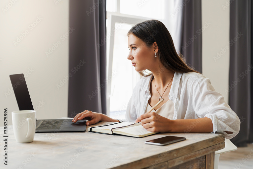 Attractive business woman sits at table in laptop, smartphone lies on table, makes notes with a pencil in notebook. Attractive business woman sits at table in laptop, smartphone lies on table, makes notes with a pencil in notebook.