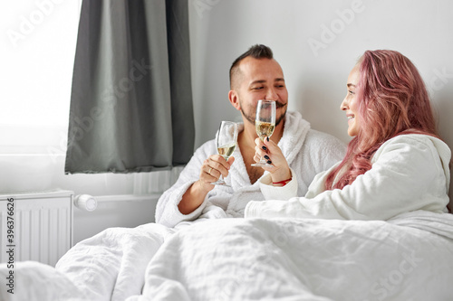 beautiful couple have romantic time on bed drinking champagne, caucasian man and woman in bathrobes drink beverage, have talk, celebrating honeymoon
