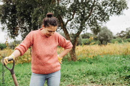 Young female farmer leaning on shovel while standing in the field and smiling. Woman working during harvesting time in autumn