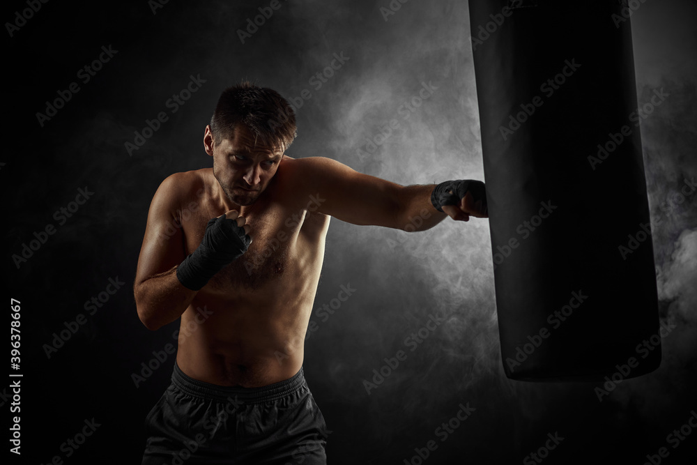 Aggressive boxer in black boxing wraps punching in boxing bag on dark background with smoke