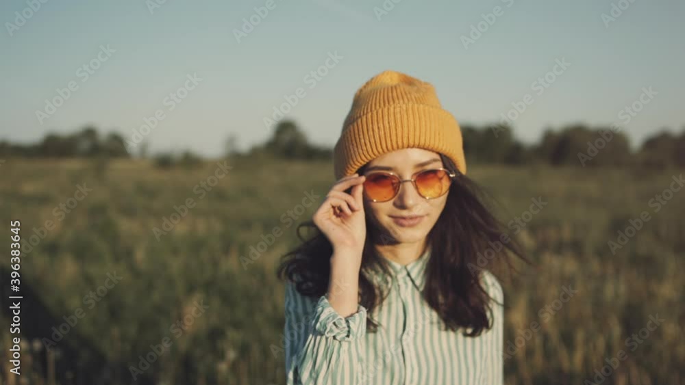 A beautiful hipster woman walking forward through a sunny field enjoying summer sunset. Stylish girl teenager in trendy sunglasses smile and walk in countryside. Concept of youth and creativity A beautiful hipster woman walking forward through a sunny field enjoying summer sunset. Stylish girl teenager in trendy sunglasses smile and walk in countryside. Concept of youth and creativity
