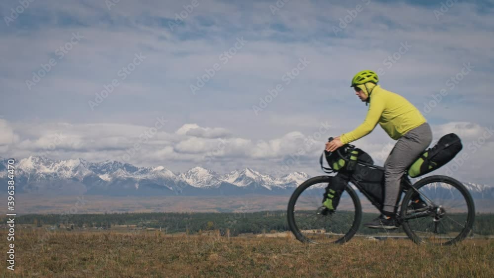 The man travel on mixed terrain cycle touring with bikepacking. The traveler journey with bicycle bags. Sport tourism bikepacking, bike, sportswear in green black colors. Mountain snow capped. The man travel on mixed terrain cycle touring with bikepacking. The traveler journey with bicycle bags. Sport tourism bikepacking, bike, sportswear in green black colors. Mountain snow capped.