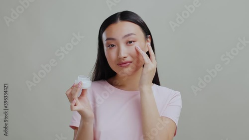Healthy looking korean woman applies moisturizing day cream holding ba jar posing isolated on grey studio background. Skincare routine. photo