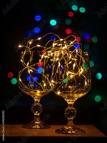 Crystal wine glasses with golden glowing garland on Christmas table bokeh light background Vertical screensaver. Romantic New Years eve dinner champagne couple in love.Magic winter holidays atmosphere