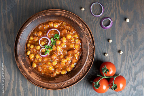 Top view of chole masala or chana local indian vegan food made of cooked chickpeas, tomatoes and cumin decorated with onion rings and parsley served in bowl on dark brown wooden background. Horizontal