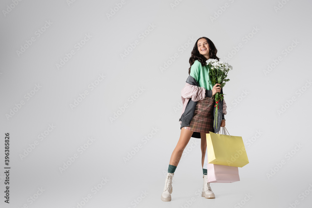 brunette young woman in stylish winter outfit posing with flowers and shopping bags on grey background brunette young woman in stylish winter outfit posing with flowers and shopping bags on grey background