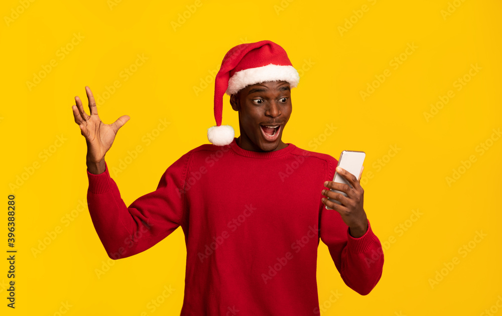 Online Christmas Sales. Surprised Black Guy In Santa Hat Looking At Smartphone Online Christmas Sales. Surprised Black Guy In Santa Hat Looking At Smartphone