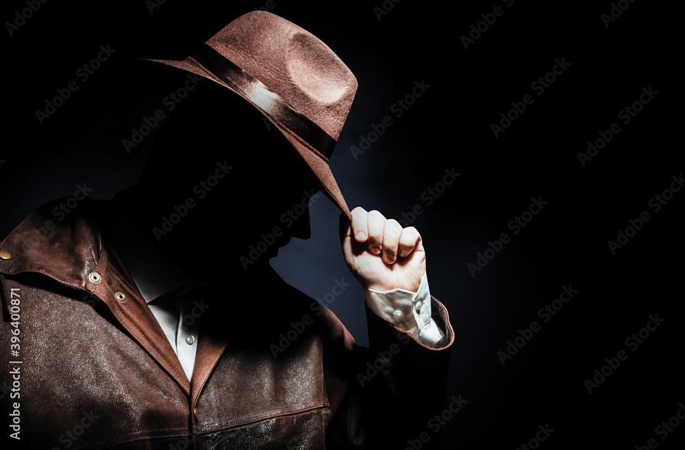 Fototapeta premium Photo of a shaded detective portrait in jacket putting on hat on black background.